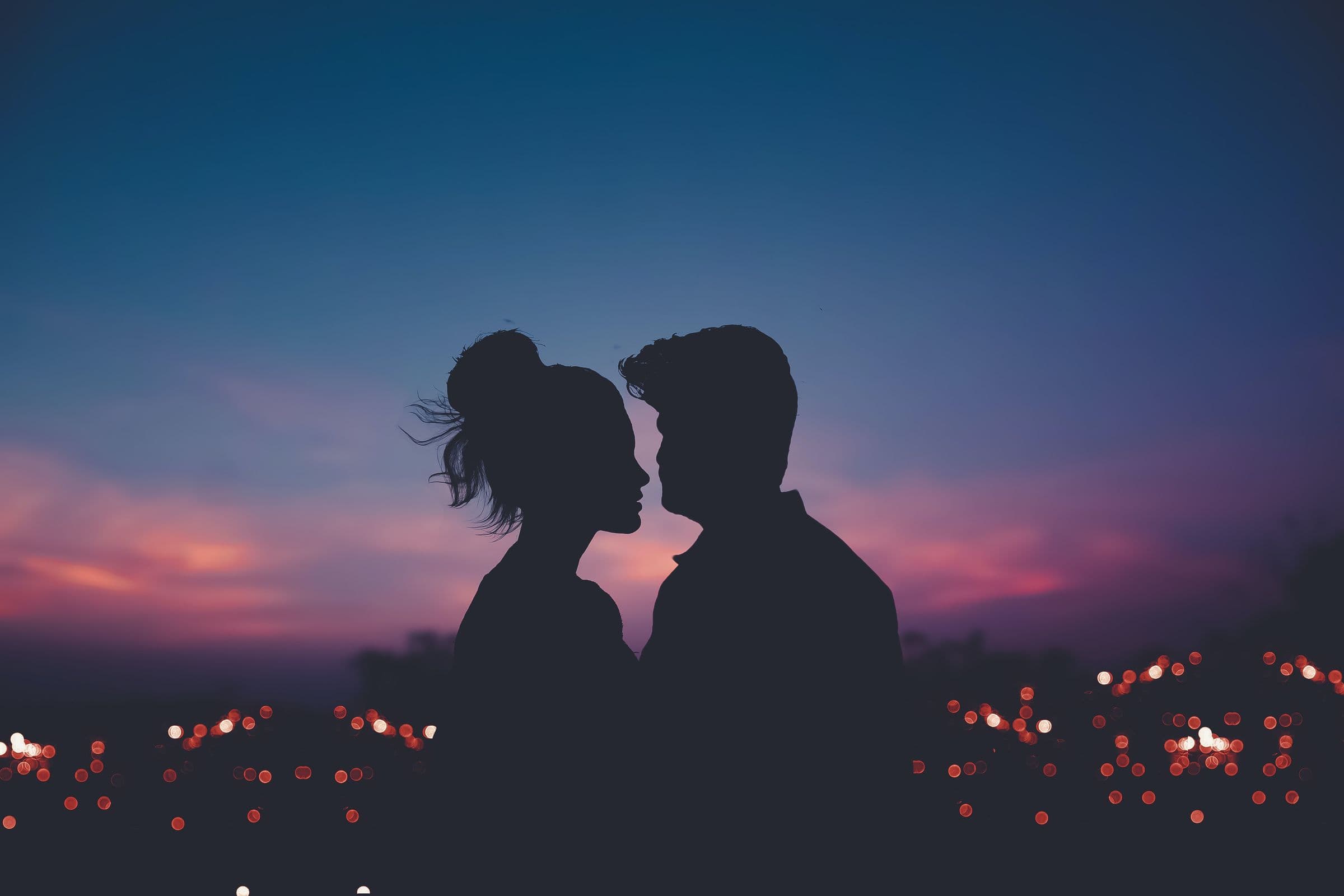 A couple in silhouette leaning in close against an indigo and pink dusk sky