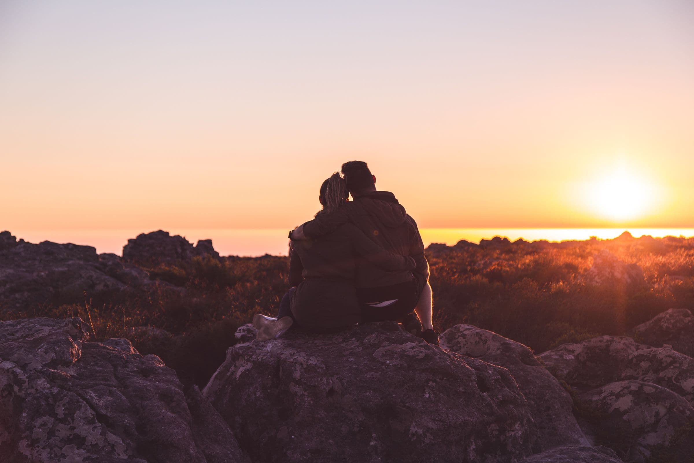 A couple sitting close together on a rocky overlook, watching the sun set